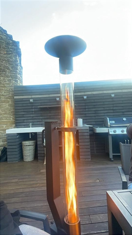 Vuurzuil op pellets met hoge vlam op een vlonder naast een buitenkeuken met gasbarbecue