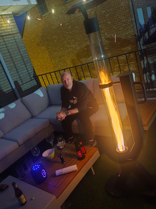 Man op een loungebank op een dakterras met lichtsnoeren en een brandende buitenhaard op pellets ernaast