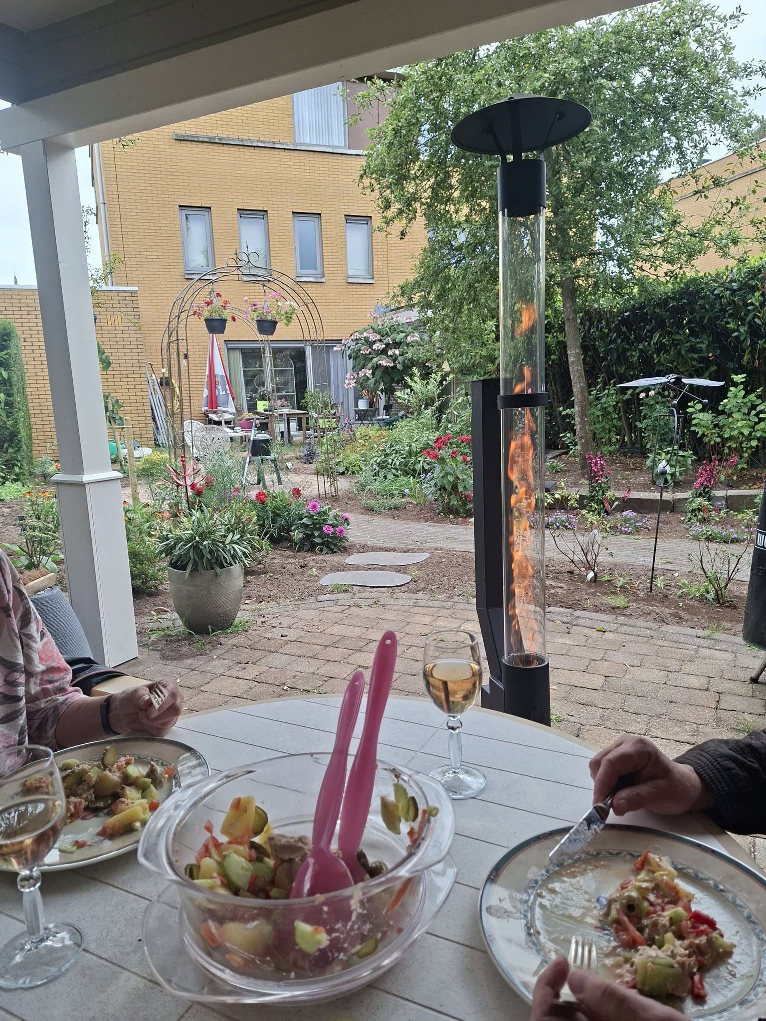 Seurue ruokailee ulkona katetun pöydän ääressä, taustalla palava pellettitakka