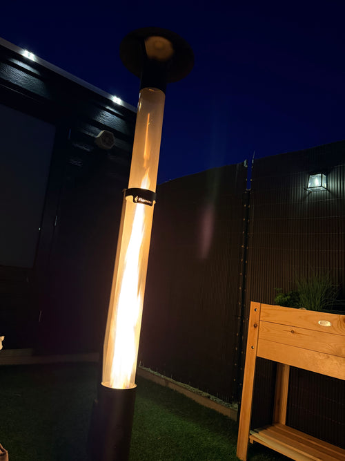 Vuurzuil op pellets brandt warm bij schemer naast een houten bank en een groene tuin