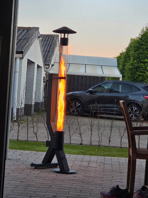 Flamesa tuinhaard op pellets brandt bij zonsondergang op een oprit naast een auto en veranda