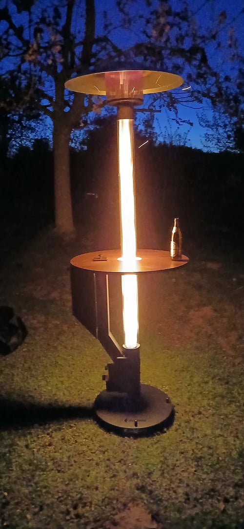 Pelletterrasverwarmer met rond tafelblad brandt in het donker op een gazon onder de bomen