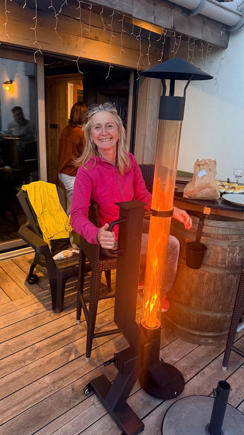Vrouw in roze trui leunt tegen een brandende terrasverwarmer op pellets op een houten vlonderterras