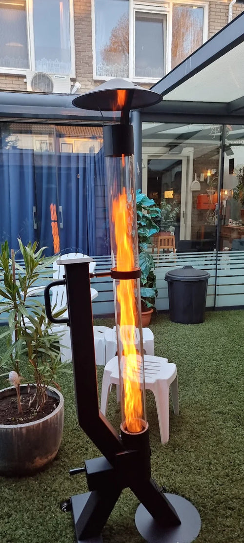 Vuurzuil op pellets brandt op kunstgras naast witte tuinstoelen bij een woning met veranda