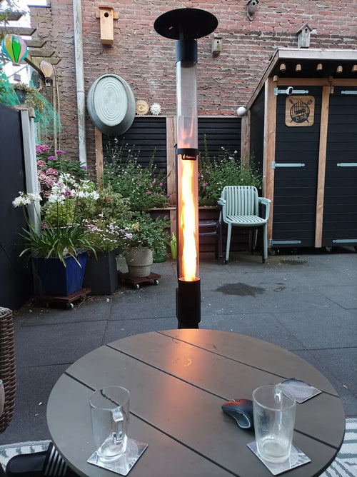 Flamesa terrasverwarmer op pellets brandt achter een ronde tuintafel met glazen in een stadstuin