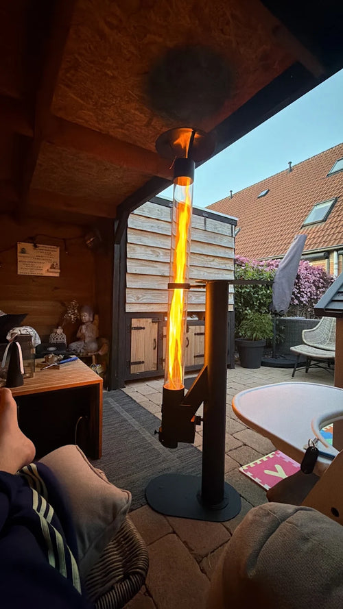 Terraskachel op pellets brandt onder een houten overkapping bij een loungestoel met plaid